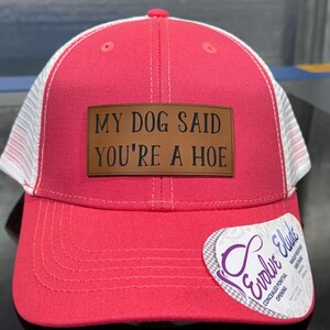 May include: A red and white trucker hat with the text "MY DOG SAID YOU'RE A HOE" on a brown leather patch. The hat has a curved brim and a mesh back. A tag on the side reads "Evolve Elude".