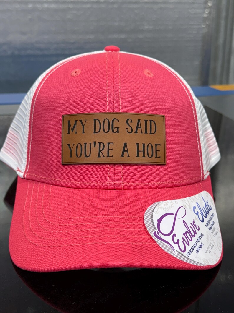 May include: A red and white trucker hat with the text "MY DOG SAID YOU'RE A HOE" on a brown leather patch. The hat has a curved brim and a mesh back. A tag on the side reads "Evolve Elude".
