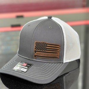 May include: A gray and white trucker hat with a brown leather patch featuring an American flag with bullets. The patch is stitched onto the front of the hat. The hat has a mesh back and a curved brim. The hat has a Richardson 112 label on the side.