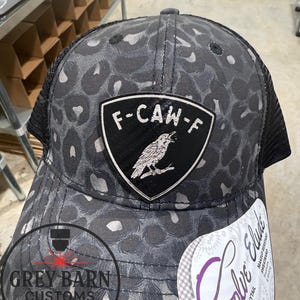 May include: A black and gray trucker hat with a leopard print pattern. The hat features a patch with the text "F-CAW-F" and a raven illustration. The hat also has a mesh back and a tag that says "Evolve Elude".