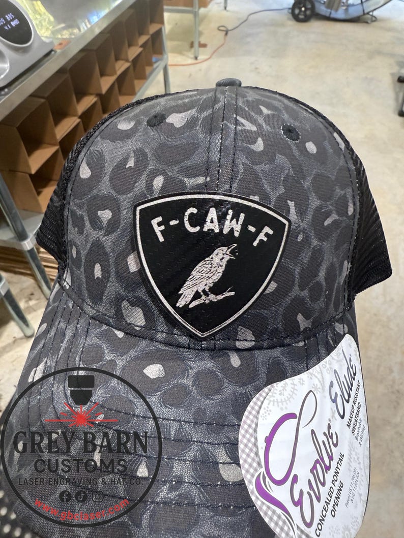 May include: A black and gray trucker hat with a leopard print pattern. The hat features a patch with the text "F-CAW-F" and a raven illustration. The hat also has a mesh back and a tag that says "Evolve Elude".