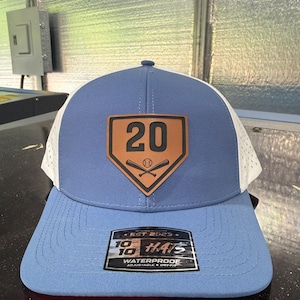 May include: A blue and white baseball cap with a brown leather patch featuring the number 20 and baseball bats. The hat has a curved brim and a mesh back. The tag reads "10/10 Hats" and "Waterproof".