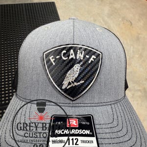 May include: A gray trucker hat with a black mesh back. The front features a patch with the text "F-CAN-F" and a bird illustration. A tag reads "RICHARDSON® ORIGINAL 112 TRUCKER ADJUSTABLE". The hat is from Grey Barn Custom.