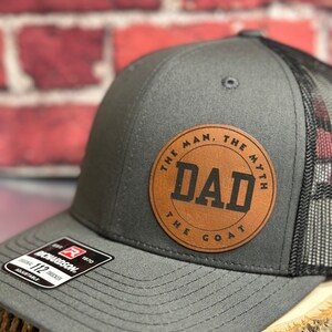 May include: A gray and black trucker hat with a brown leather patch that says "The Man, The Myth, Dad, The Goat".
