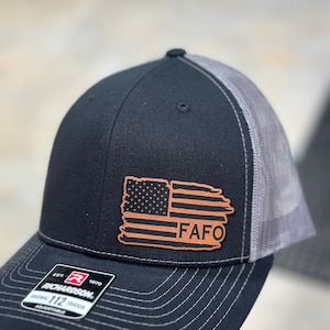 May include: Black and grey trucker hat with a brown leather patch featuring an American flag and the text "FAFO". The hat is a Richardson 112 adjustable trucker hat.