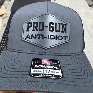 May include: Gray trucker hat with a black mesh back and a silver emblem that reads "PRO-GUN ANTI-IDIOT." The hat has a curved brim and an adjustable strap. The Richardson brand tag is visible.