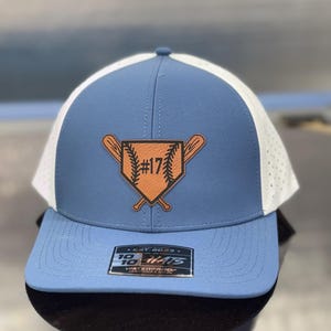 May include: A blue and white baseball cap with a leather patch featuring a baseball diamond and crossed bats. The number "17" is visible on the patch. The hat has a curved brim and a mesh back.