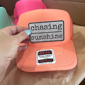 May include: An orange trucker hat with a mesh back and a curved brim. The hat features a rectangular patch with the words "chasing sunshine" in black. A white label with the brand name "OTTO" is also visible.