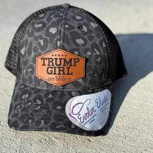 May include: A black and grey trucker cap with a leopard print pattern. It has a brown leather patch with the text "TRUMP GIRL GET OVER IT". The cap features a mesh back and a curved peak.