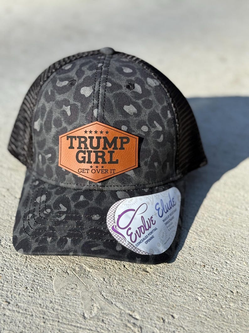 May include: A black and grey trucker cap with a leopard print pattern. It has a brown leather patch with the text "TRUMP GIRL GET OVER IT". The cap features a mesh back and a curved peak.