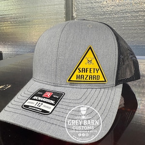 May include: Grey Richardson 112 trucker cap with a black mesh back. The front panel has a yellow triangle with a skull and crossbones and the words "SAFETY HAZARD". The cap is adjustable.