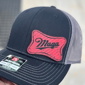 May include: A black and grey trucker hat with a red patch that says "Maga" in black lettering. The hat has a black brim and a grey mesh back.