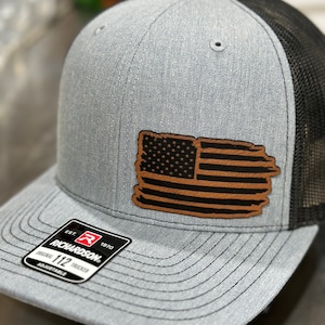 Peut inclure: Un chapeau de camionneur gris et noir avec un patch en cuir marron représentant le drapeau américain. Le chapeau a un dos en maille noire et un devant gris avec une étiquette Richardson 112.