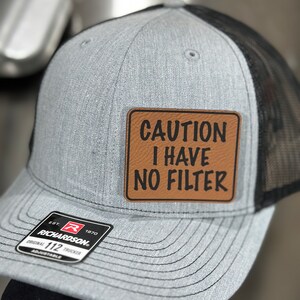 May include: A gray and black trucker hat with a brown leather patch that says "CAUTION I HAVE NO FILTER".