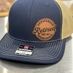 May include: A navy blue and tan trucker hat with a brown leather patch that reads "Officially Retired Not My Problem Anymore". The hat has a Richardson logo and the text "Est. 1970 Original 112 Trucker Adjustable".