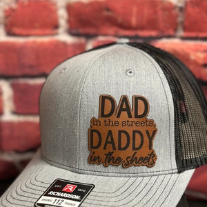 May include: A gray and black trucker hat with a brown leather patch that reads "DAD in the streets, DADDY in the sheets". The hat has a black mesh back and a gray front with a Richardson logo.