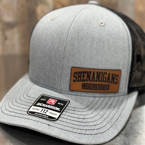 May include: A grey and black trucker hat with a brown leather patch that says "Shenanigans Coordinator" and a black and red Richardson 112 label.