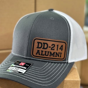 May include: A grey and white trucker hat with a brown leather patch that says "DD-214 ALUMNI". The hat is adjustable and has a Richardson 112 label.