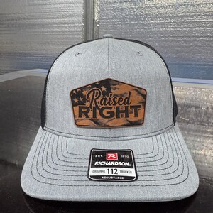 May include: Gray trucker hat with a black mesh back and a curved brim. The front features a brown leather patch with the words "Raised RIGHT" and a flag design. The hat has a Richardson tag that says "Original 112 Trucker".