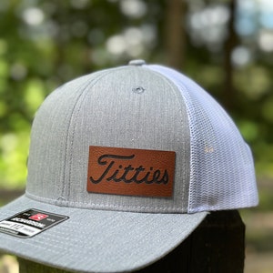 May include: A gray and white trucker hat with a brown leather patch that says "Titties" in a cursive font.