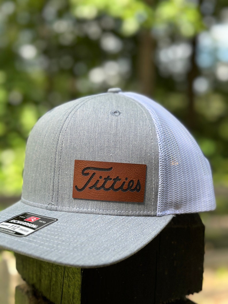 May include: A gray and white trucker hat with a brown leather patch that says "Titties" in a cursive font.