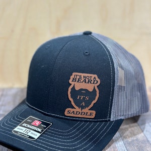 May include: A black and gray trucker hat with a brown leather patch that says "It's not a beard, it's a saddle".