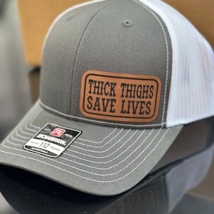 May include: Gray and white trucker hat with a brown leather patch that says "Thick Thighs Save Lives".
