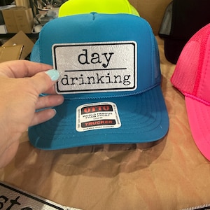 May include: A turquoise trucker hat with a white rectangular patch that reads "day drinking" in black text. The hat has a mesh back and a curved brim. Other hats in neon yellow and pink are visible.