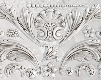 May include: White decorative relief with ornate floral and leaf designs. The relief features a central shell-like element, symmetrical swirling leaves, and small flower accents. The design is detailed and textured.