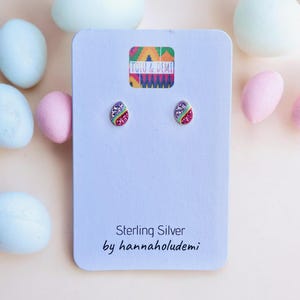 May include: Sterling silver Easter egg stud earrings with colourful glitter accents. The earrings are displayed on a light blue card with the text "Tolu & Demi" and "Sterling Silver by hannaholudemi". Pastel Easter eggs surround the card.