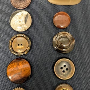 May include: A collection of various buttons in different shapes, sizes, and materials. The buttons include brown, gold, and iridescent colors. Some buttons have two or four holes, while others have decorative designs.