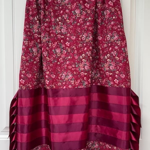 Op de afbeelding: Een rok tot op de knie met een donkerrode bloemenprint op het bovenste en onderste gedeelte. Het middengedeelte bestaat uit horizontale banen satijnlint in afwisselende tinten magenta en bordeauxrood, afgewerkt met geplooide lintranden.