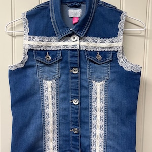 May include: A blue denim vest with white lace trim around the collar, armholes, and down the front. The vest has a button closure and two pockets.