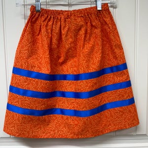 May include: An orange skirt with a floral pattern and three horizontal blue stripes. The skirt is hanging on a clear hanger against a white background. The skirt is made of fabric.