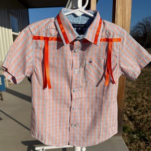 May include: A short-sleeved, button-down shirt in a plaid pattern of orange and light blue. The shirt has bright orange ribbons attached to the collar and shoulders. The shirt is size 2T and has a small pocket on the chest.