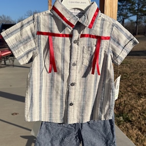 May include: A two-piece Calvin Klein Jeans outfit for a child. The short-sleeved shirt is striped in white, blue, and gray, with red ribbon accents. The matching shorts are a solid blue. The outfit is displayed on a white hanger.