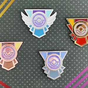 May include: Four enamel pins with a Poké Ball design in different colors and styles. The pins are silver, gold, rose gold, and blue.