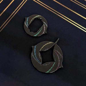 May include: Two black metal pins with a unique design. Each pin features a circular shape with a stylized leaf design and iridescent glitter accents. The pins are displayed on a dark blue surface with gold lines.