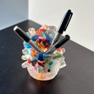 Puede incluir: Un porta lápices de plástico transparente lleno de marcadores y bolígrafos de colores. El porta lápices tiene la forma de un racimo de pequeñas bolas de colores.