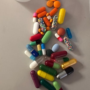 May include: Assorted pills and capsules in various colors and shapes, including orange, white, green, and blue. Some capsules are clear with colorful granules inside. A small white plastic cup is in the upper left corner.