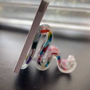 May include: A white book stand with a clear, wavy design. The stand is decorated with colourful confetti-like pieces. The stand is holding a white book.