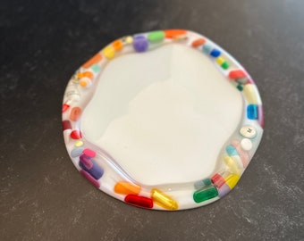 Puede incluir: Una bandeja circular blanca de resina con bordes ondulados, que presenta un borde incrustado con varias cápsulas de pastillas coloridas de diversas formas y pequeñas tabletas redondas en tonos rojo, naranja, amarillo, verde, azul, morado y blanco.