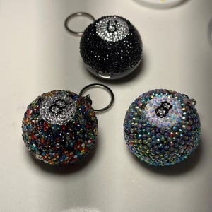 Könnte beinhalten: Drei Schlüsselanhänger in Form einer 8-Ball mit unterschiedlichen Designs. Einer ist schwarz mit einer weißen 8, einer ist regenbogenfarben mit einer weißen 8 und einer ist schillernd mit einer weißen 8.