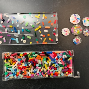 May include: A rectangular clear resin tray containing scattered multi-colored pill capsules, a smaller rectangular tray densely packed with various colorful capsules, and five small circular resin coasters embedded with assorted pill capsules.