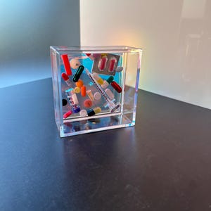 May include: A clear plastic cube filled with a variety of colorful pills and a syringe. The cube is sitting on a dark surface.