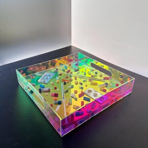 May include: A square iridescent tray filled with various colorful pills, capsules, and syringes. The tray has a handle on one side and is sitting on a black surface.