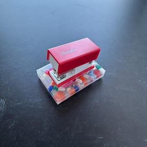 May include: A red Swingline stapler with a clear plastic tray filled with colorful paper clips, push pins, and staples.