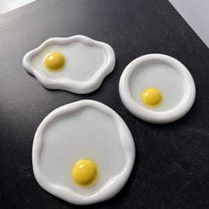 May include: Three white ceramic dishes shaped like fried eggs with yellow yolks. The dishes are on a black surface.