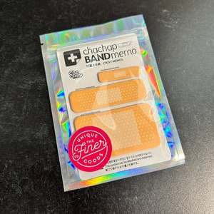 May include: A pack of adhesive memo notes shaped like band-aids. The pack includes three sizes of notes: 1.0 x 4.0 cm, 2.0 x 7.0 cm, and 4.0 x 10.0 cm. The notes are beige with white polka dots and a white border. The packaging is silver with a holographic finish and features the text "chachap BAND memo" and "付属メモ紙 STICKY MEMOS".