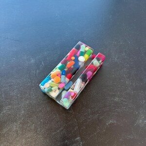 Könnte beinhalten: Ein klarer Telefonständer aus Harz, gefüllt mit bunten Pillen und Kapseln. Der Ständer ist rechteckig mit einem Schlitz zur Aufnahme eines Telefons. Die Pillen sind in verschiedenen Farben, darunter Orange, Blau, Grün und Rosa.